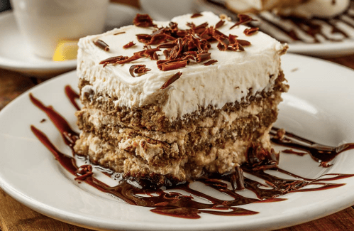 A slice of layered dessert with cream frosting, chocolate shavings, and a drizzle of chocolate sauce on a white plate.