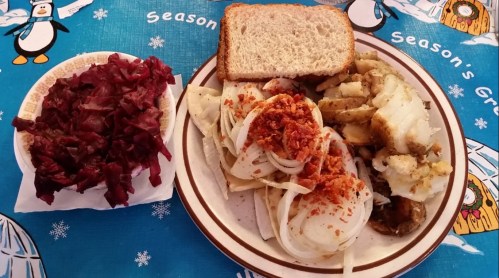 A plate with sliced potatoes, onions, and a topping, alongside a bowl of red cabbage and a slice of bread.