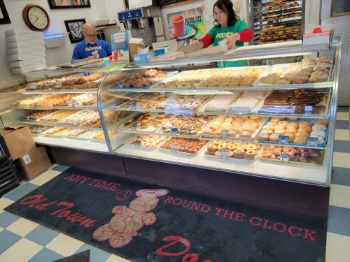 A display case filled with various donuts and pastries in a bakery, with staff members in colorful shirts behind the counter.