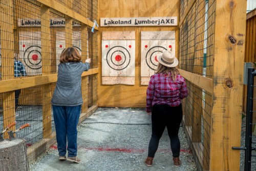 Two women at an axe-throwing venue, one preparing to throw while the other watches, with targets in the background.
