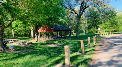 A red picnic shelter surrounded by green trees and grass, with picnic tables nearby in a park setting.