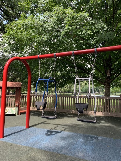 Three swings hang from a red frame in a playground, surrounded by green trees and a wooden fence.