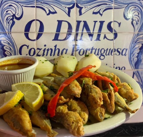 A plate of fried fish with lemon, red pepper, and potatoes, served with a dipping sauce against a tiled background.