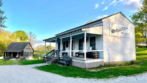 A historic building labeled "Shields" with a porch, surrounded by green grass and trees, next to a smaller structure.