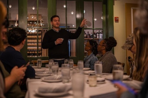 A man speaks to a group at a dinner table, with glasses of water and plates set, in a cozy restaurant setting.