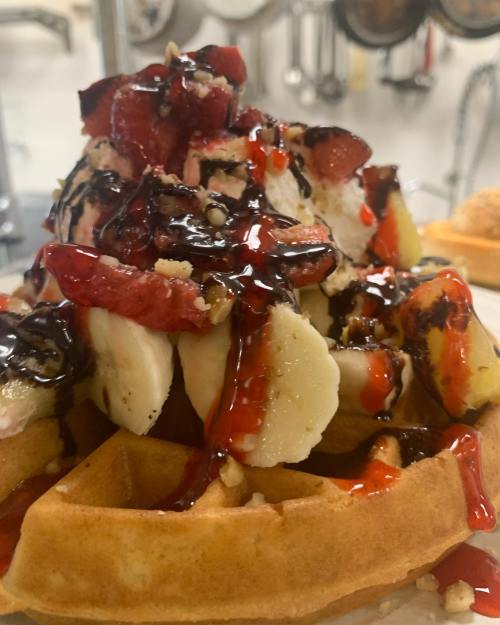 A close-up of a waffle topped with bananas, strawberries, chocolate syrup, and a drizzle of red sauce.