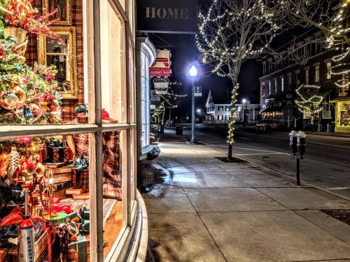 A festive storefront with a decorated Christmas tree, illuminated by streetlights on a quiet, decorated street at night.