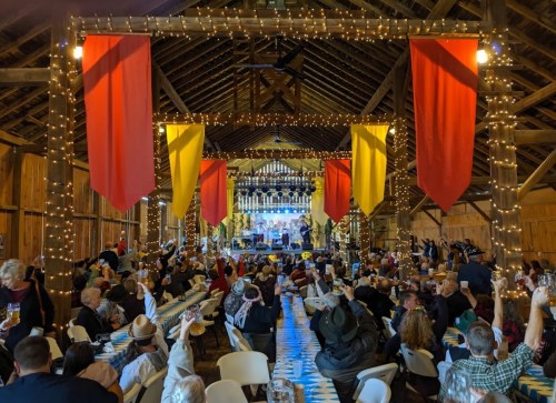 A lively barn event with guests enjoying dinner, colorful banners, string lights, and a band performing on stage.