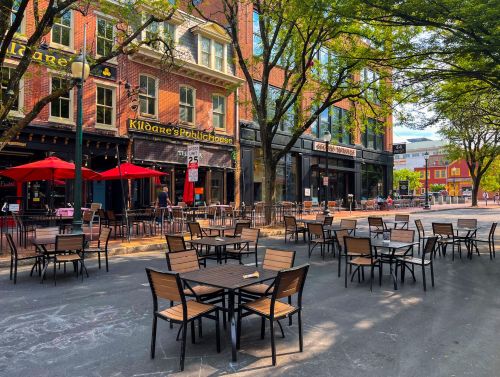 Outdoor dining area with empty tables and red umbrellas, surrounded by trees and buildings in a lively urban setting.