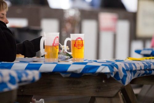Two mugs of beer sit on a blue and white checkered tablecloth, with a person partially visible in the background.