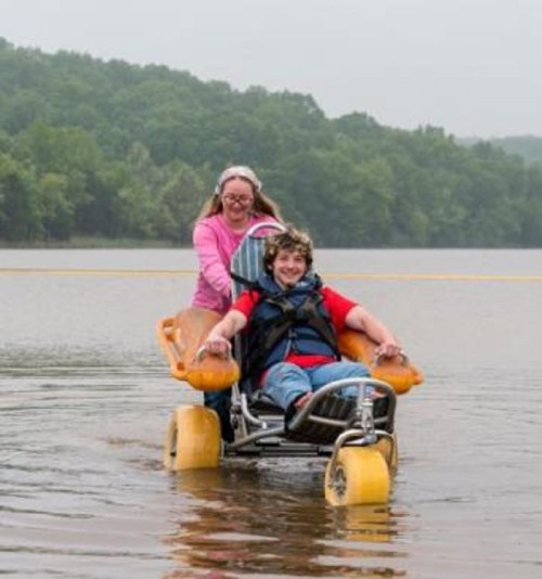 A child in a wheelchair is being pushed through shallow water by an adult, surrounded by trees and a cloudy sky.