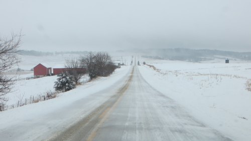 A snowy rural road stretches into the distance, flanked by fields and a red barn under a gray sky.