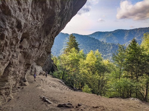 A scenic view of a rocky cave opening, surrounded by lush green trees and mountains under a partly cloudy sky.