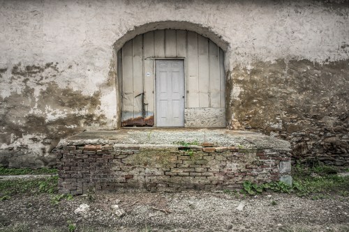 A weathered wall with a simple door set on a stone platform, surrounded by peeling paint and overgrown greenery.