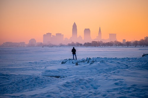 A person stands on snowy ground, gazing at a city skyline during a colorful sunrise.