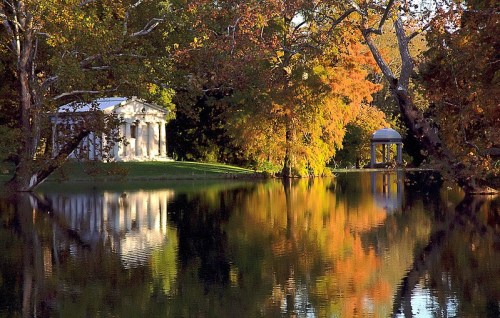 A serene landscape featuring a pond reflecting colorful autumn trees and classical structures on the shore.