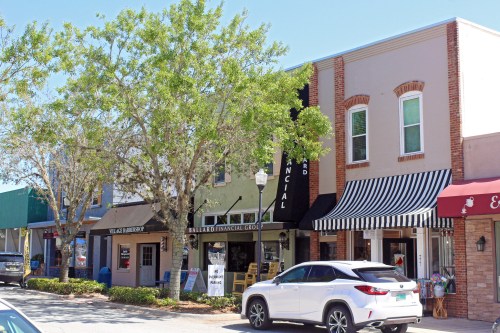 A sunny street scene featuring shops with awnings, trees, and a parked car.