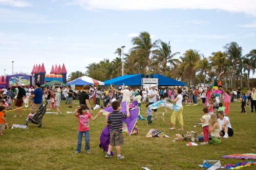 A lively outdoor festival scene with families, children flying kites, and colorful tents under a clear blue sky.