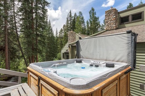 A hot tub on a wooden deck surrounded by tall trees and a house, with a cloudy blue sky in the background.