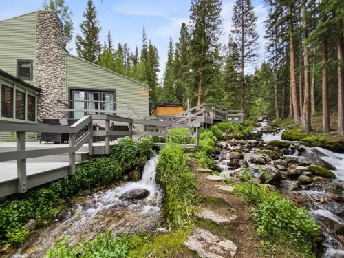 A serene landscape featuring a wooden deck beside a flowing stream, surrounded by lush greenery and tall trees.