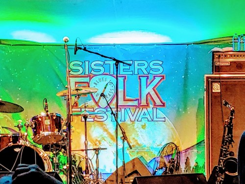 Stage setup for the Sisters Folk Festival, featuring drums, a saxophone, and a colorful backdrop.