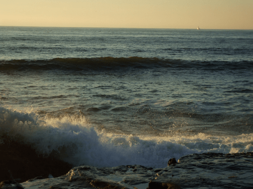 Waves gently crash on rocky shore at sunset, with a distant boat on the horizon under a golden sky.