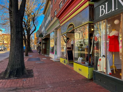 A charming street scene featuring colorful storefronts, a red dress in a window, and trees lining the sidewalk.