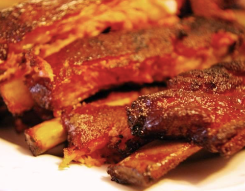 Close-up of succulent, glazed barbecue ribs with a smoky, caramelized crust on a white plate.