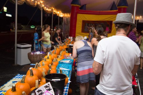 A festive scene with people at a pumpkin-themed event, featuring decorated tables and a bounce house in the background.