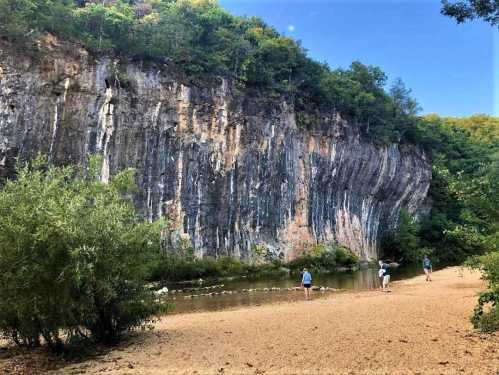 A sandy riverbank with people walking near a tall, rocky cliff surrounded by lush greenery under a clear blue sky.
