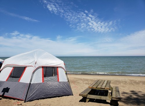 A tent on a sandy beach with a picnic table, overlooking a calm lake under a blue sky with scattered clouds.