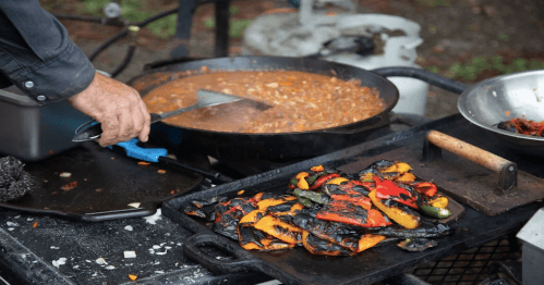 A hand stirs a pot of beans while grilled vegetables sit on a nearby grill.