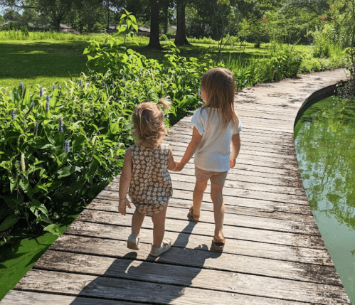 Two young children walk hand in hand along a wooden path beside a green pond, surrounded by lush greenery.