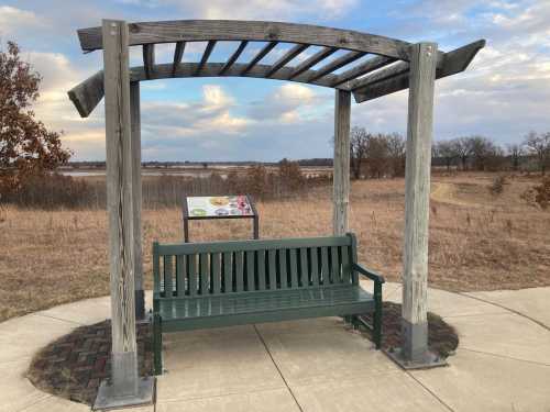 A green bench under a wooden pergola, with a scenic view of a field and water in the background.