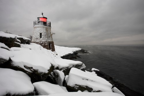 A snow-covered lighthouse stands on rocky shore under a cloudy sky, with a red light glowing at the top.