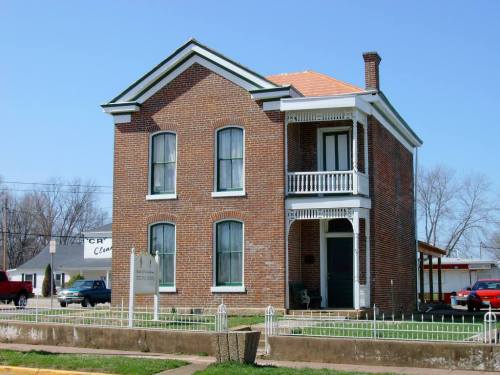 Historic brick house with a porch, white trim, and a sign in front, set against a clear blue sky.