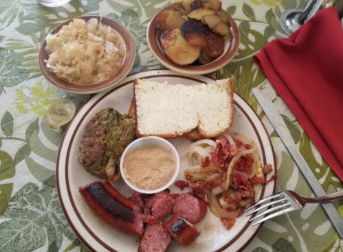 A plate of sausage, sauerkraut, onions, and a slice of bread, with a side of potatoes on a floral tablecloth.