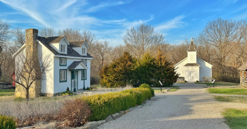 A scenic view of a white house and a small church surrounded by trees and a gravel path under a blue sky.