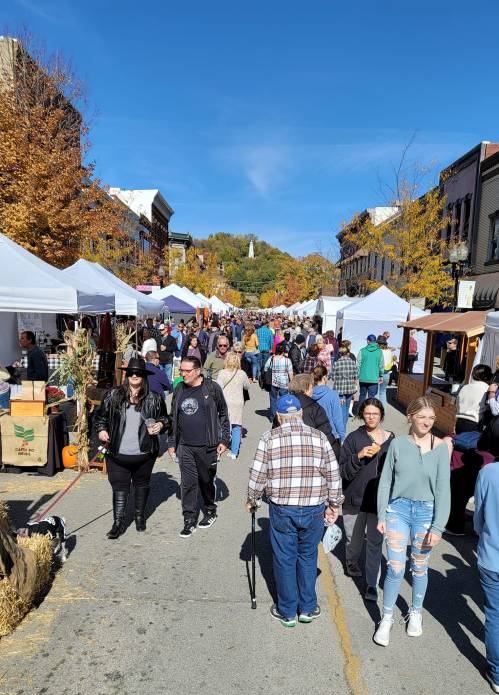 A bustling street market with people browsing tents, surrounded by autumn trees and clear blue skies.