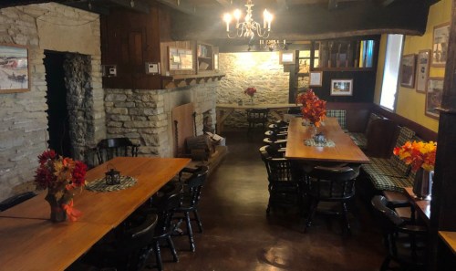 Cozy dining area with wooden tables, chairs, and autumn decorations, featuring a stone fireplace and warm lighting.