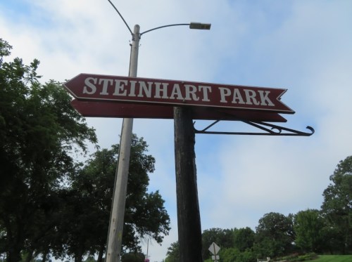 Signpost pointing to Steinhart Park, surrounded by trees and a cloudy sky.