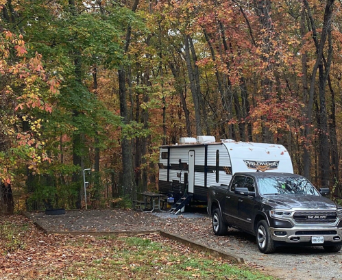 A black truck parked next to a travel trailer in a wooded area with colorful autumn leaves.