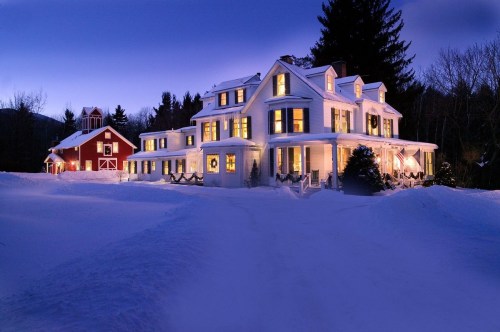 A charming white house and red barn illuminated at dusk, surrounded by snow-covered ground and trees.