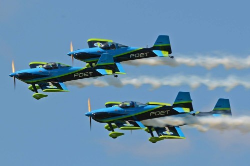 Three blue and green stunt planes flying in formation, leaving trails of smoke against a clear blue sky.