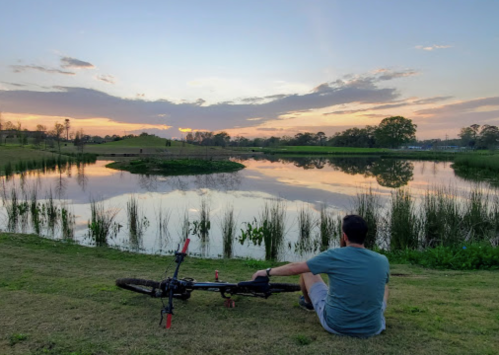 A person sits by a calm lake at sunset, with a bicycle resting beside them and reflections on the water.