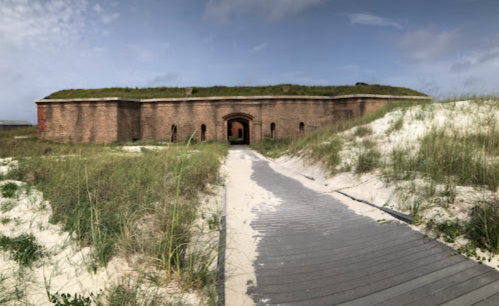 Historic brick fort surrounded by grass and sand, with a wooden pathway leading to its entrance.