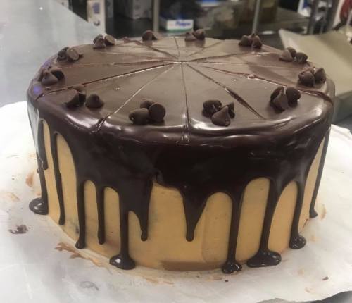 A round chocolate cake with a glossy ganache topping and chocolate chips, featuring drips down the sides.