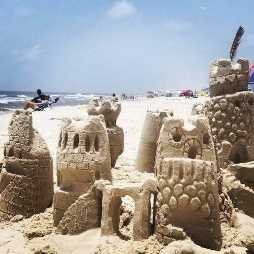 Sandcastle structures on a beach, with people relaxing in the background and waves in the distance.