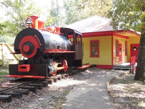A vintage black and red steam locomotive next to a bright yellow train station surrounded by trees.