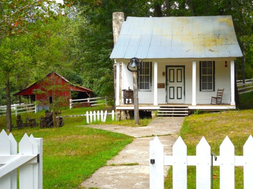 A quaint white house with a metal roof, surrounded by greenery and a red barn in the background, with a white picket fence.